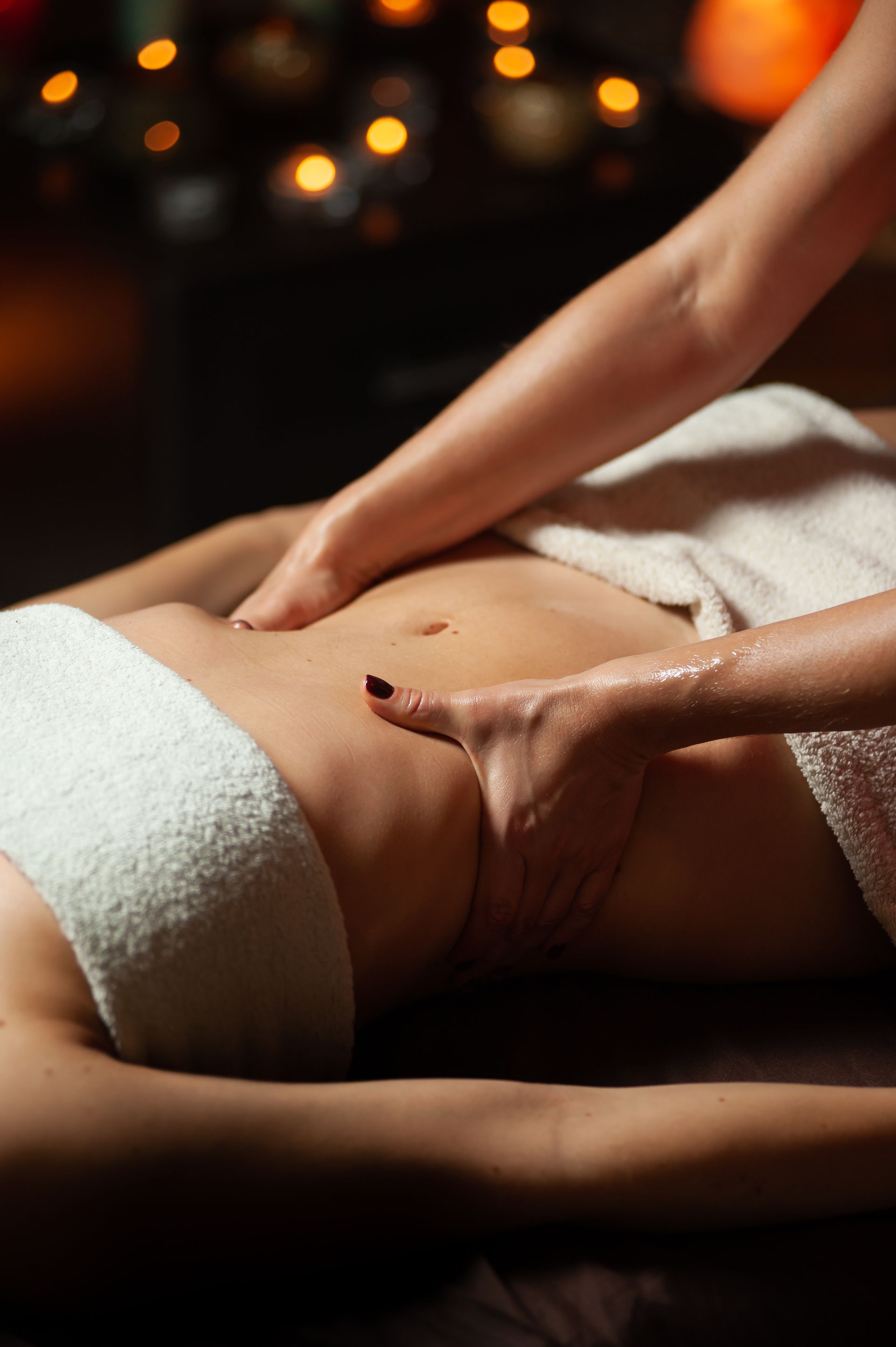 Manos masajeando el abdomen de una persona, cubierta con una toalla blanca, en un spa con luz tenue.
