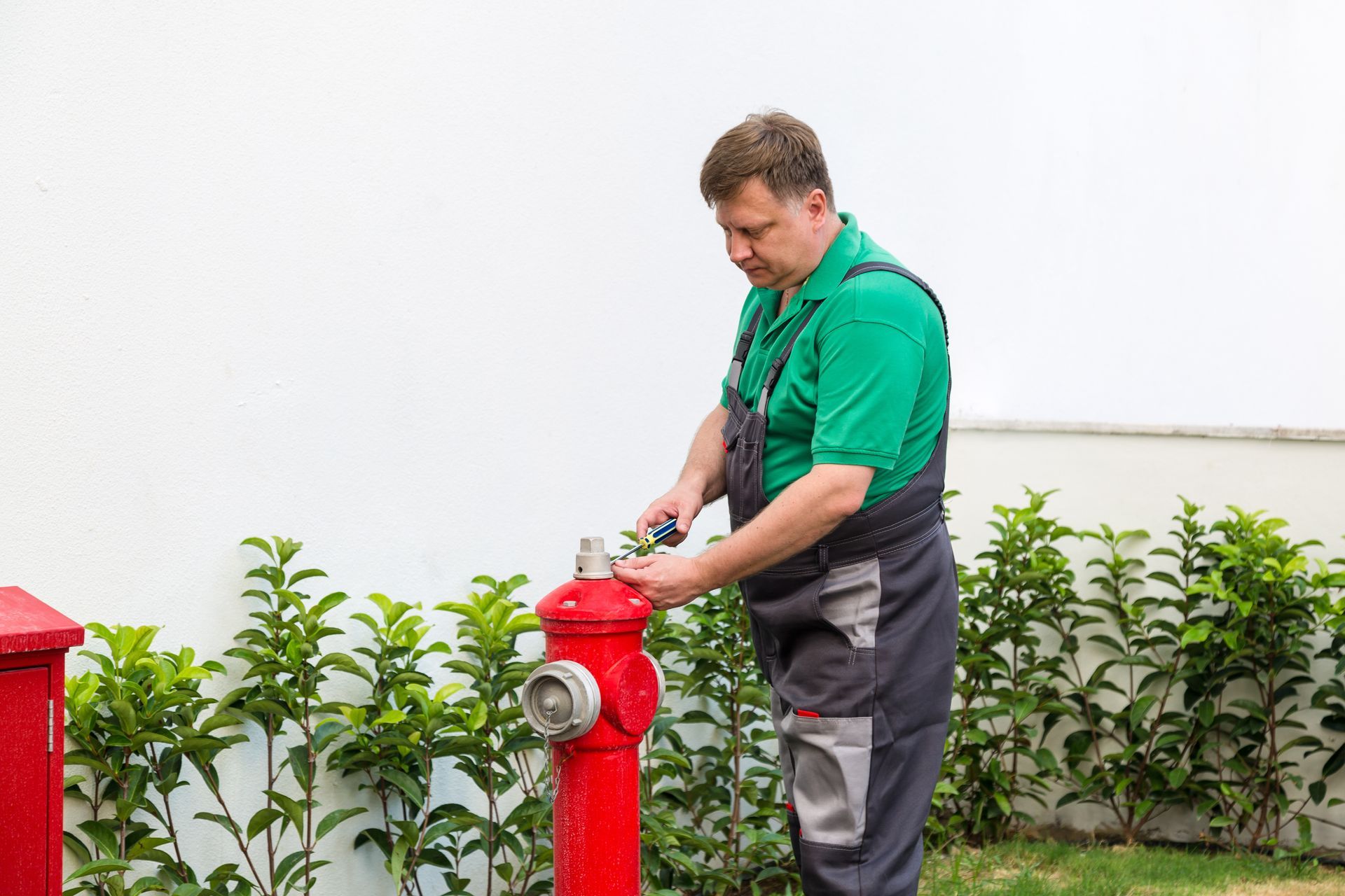 A worker in grey overalls and a green shirt is performing a fire hydrant inspection at a property.