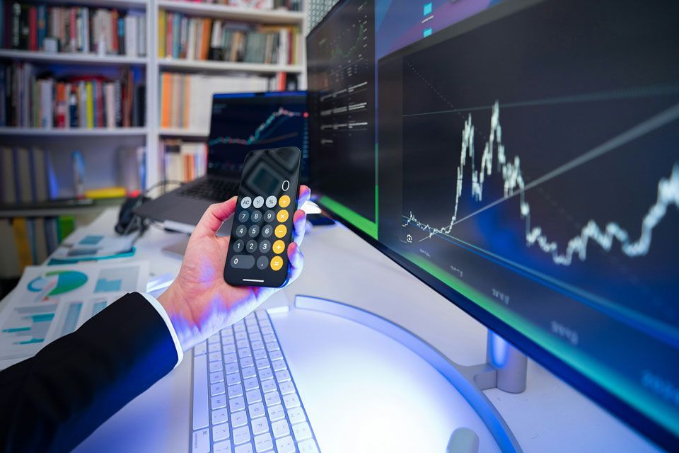 Person holding calculator, monitoring stock charts on multiple computer screens.