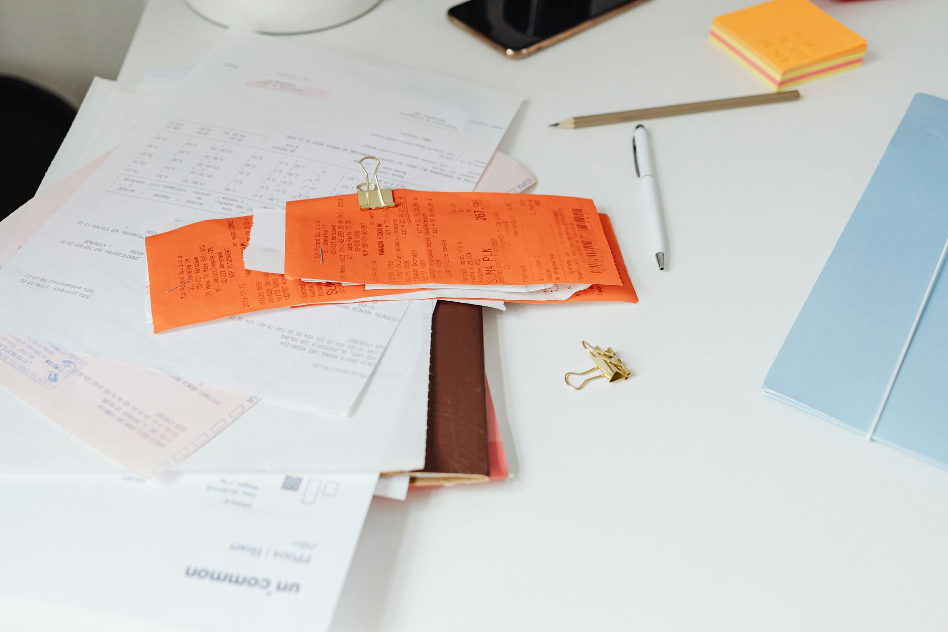 White desk with scattered papers, orange file clipped, pen, pencil, sticky notes, and phone.
