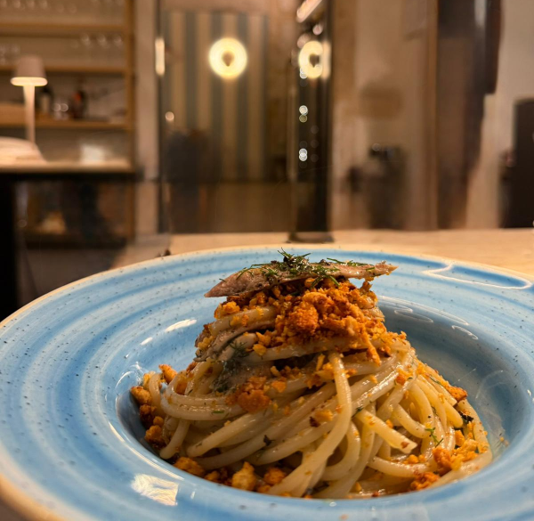 Piatto di pasta con pangrattato, erbe aromatiche e pesce su un piatto blu, in un ambiente da ristorante.
