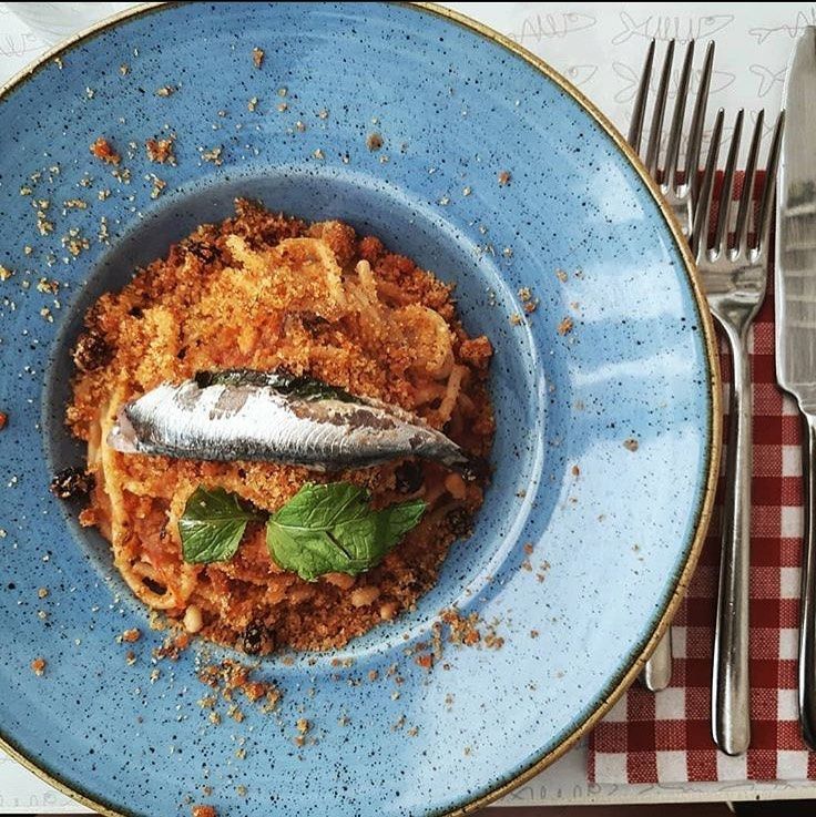 Piatto di spaghetti con pangrattato, pesce e basilico su un piatto blu. Tavola apparecchiata con posate e tovagliolo a quadretti.