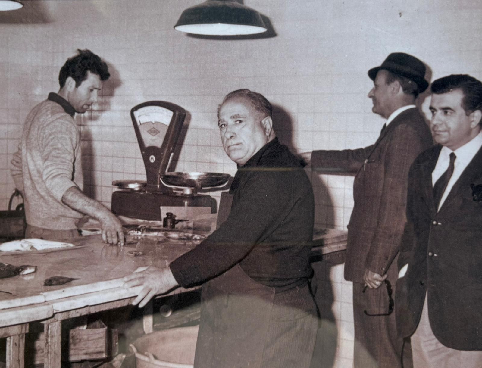 Macelleria con tre uomini in giacca e cravatta e un macellaio, bilancia d'epoca, pareti piastrellate.