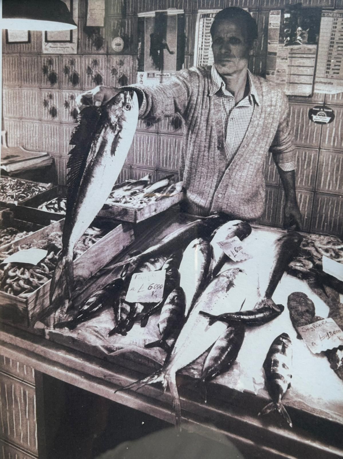Un pescivendolo tiene in mano un grosso pesce, in piedi dietro un bancone che espone vari frutti di mare in un mercato.