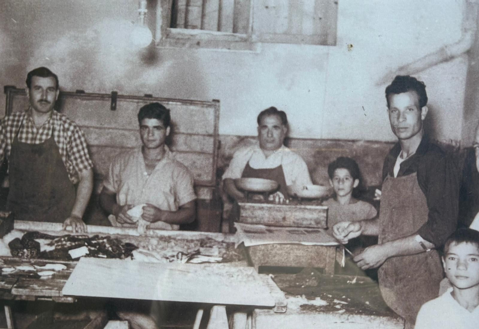 Un gruppo di persone, forse una famiglia, che lavora a un tavolo, probabilmente in un laboratorio. Tutti indossano un grembiule.