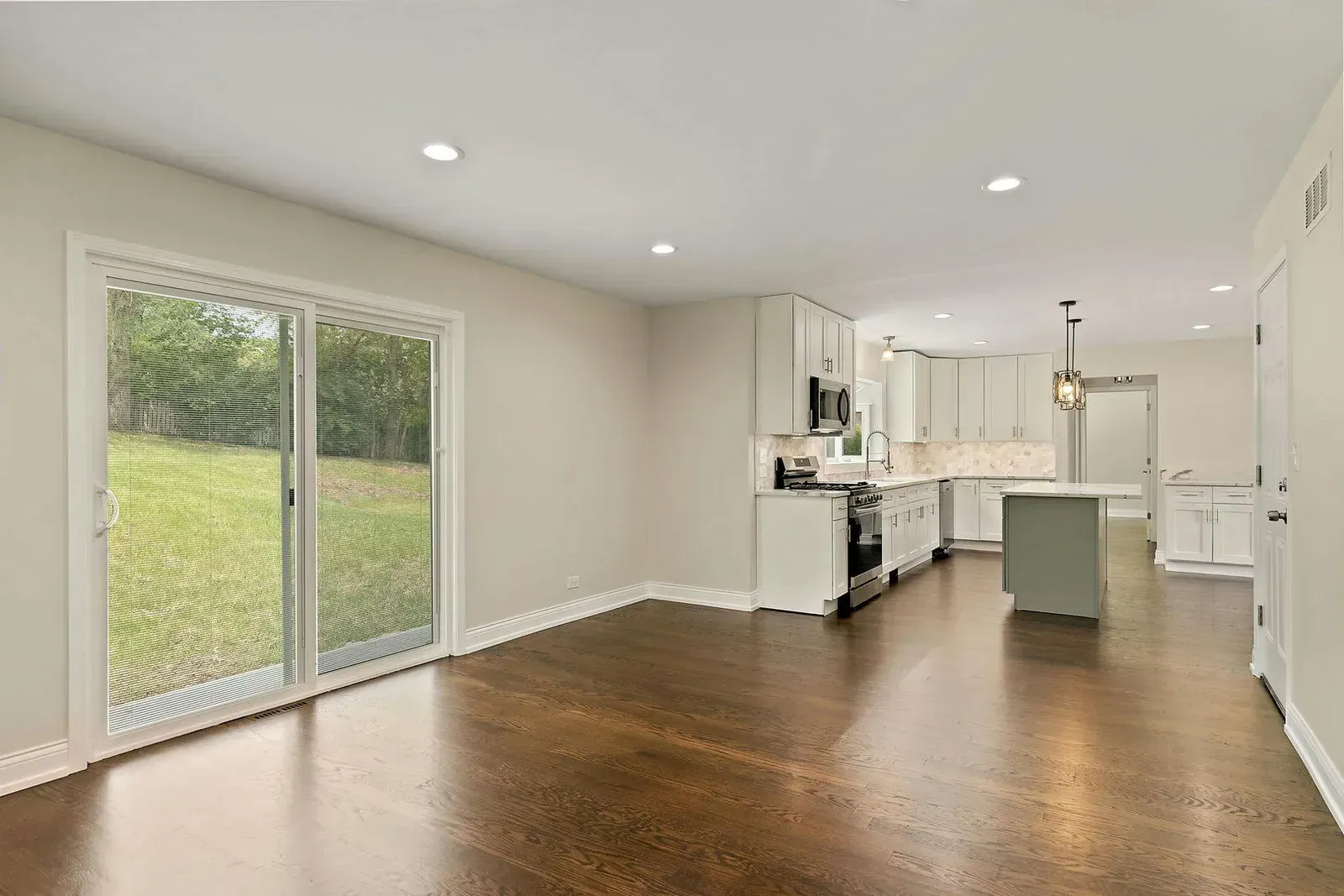 An empty living room with hardwood floors and sliding glass doors leading to a kitchen.