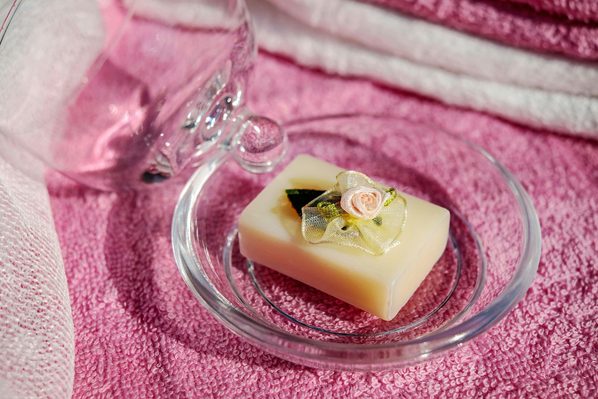 Soap bar with floral accents on a glass dish, beside a clear glass, and pink towel background.