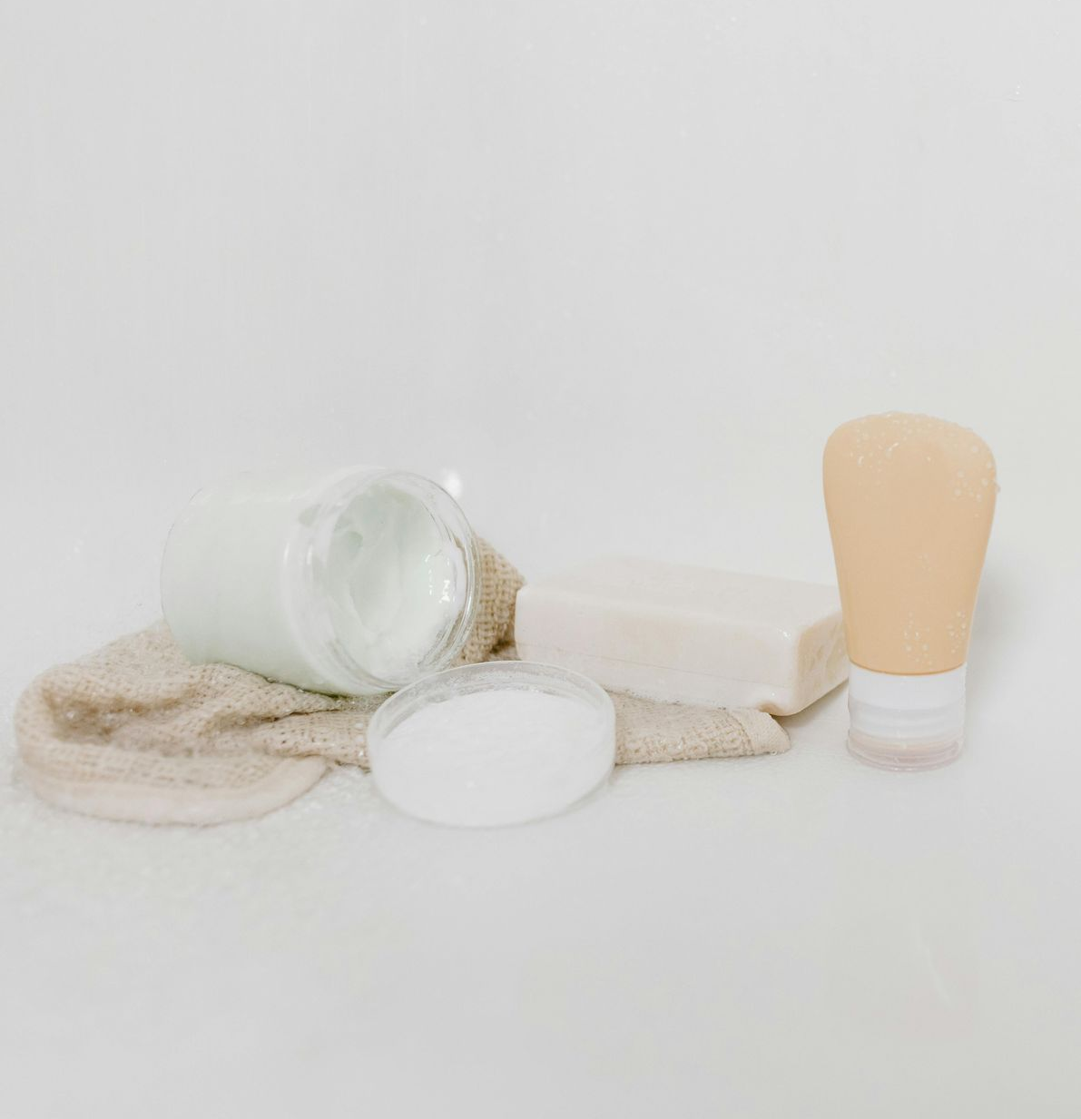 White bathroom products: jar, soap bar, lotion tube, and sponge, on a white surface.
