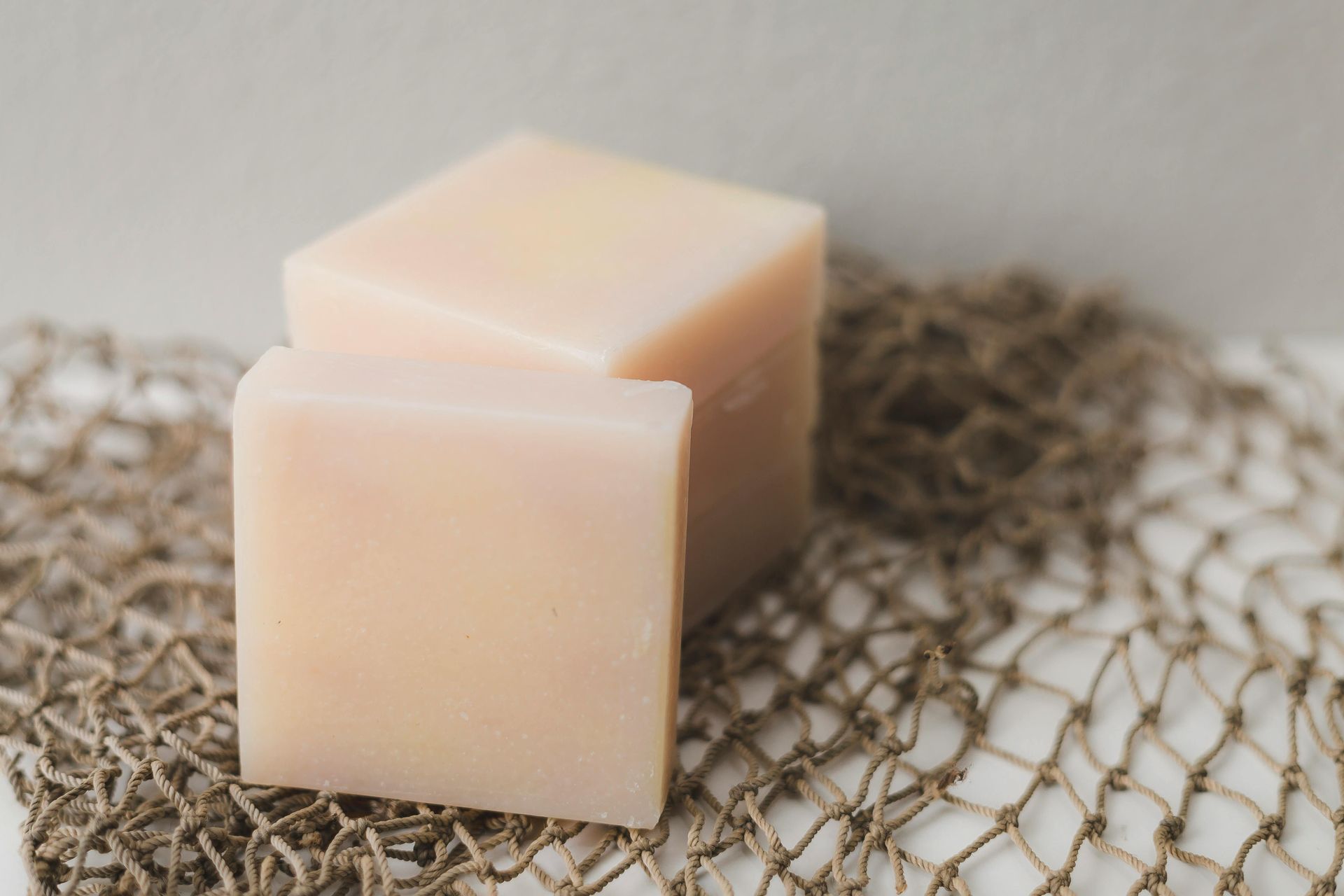 Two square, pale pink soap bars on a beige netting background.