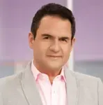 Un hombre con traje y camisa rosa está parado frente a una pared violeta.