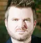 Un hombre con barba mira a la cámara con una mirada seria en su rostro.