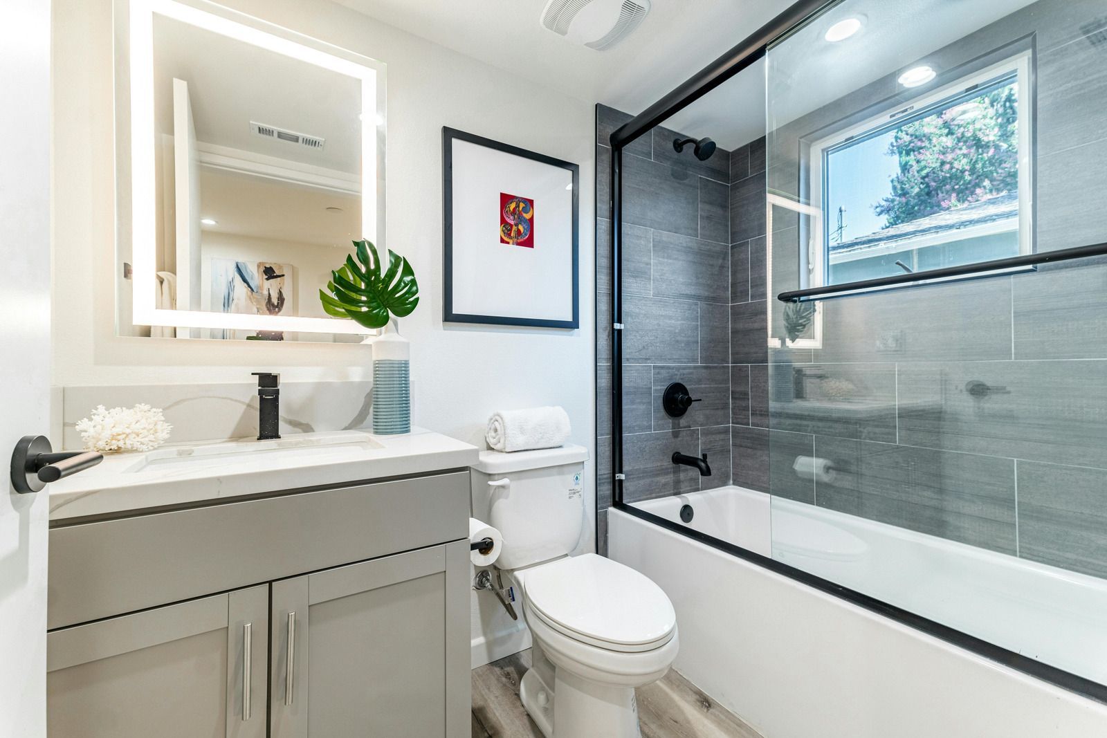 Modern bathroom with white vanity, lit mirror, toilet, and glass-enclosed bathtub shower.
