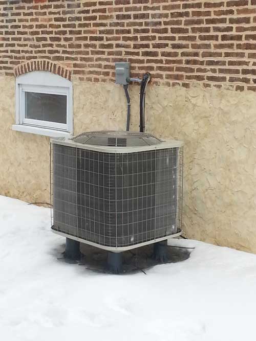 An air conditioner is sitting outside of a brick building in the snow.