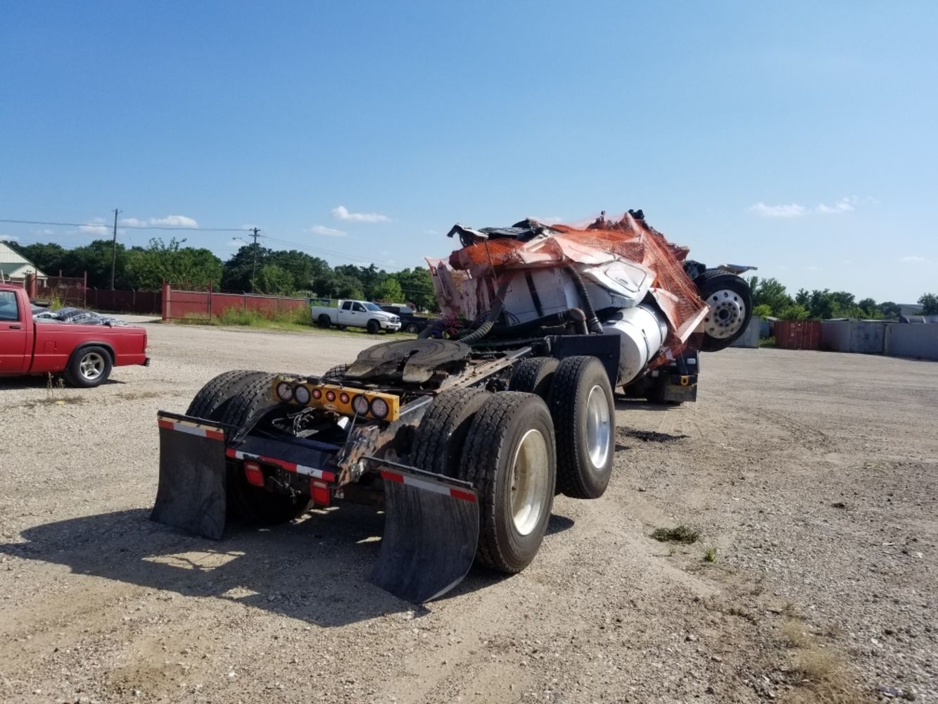 A red truck is parked next to a wrecked semi truck in a gravel lot.