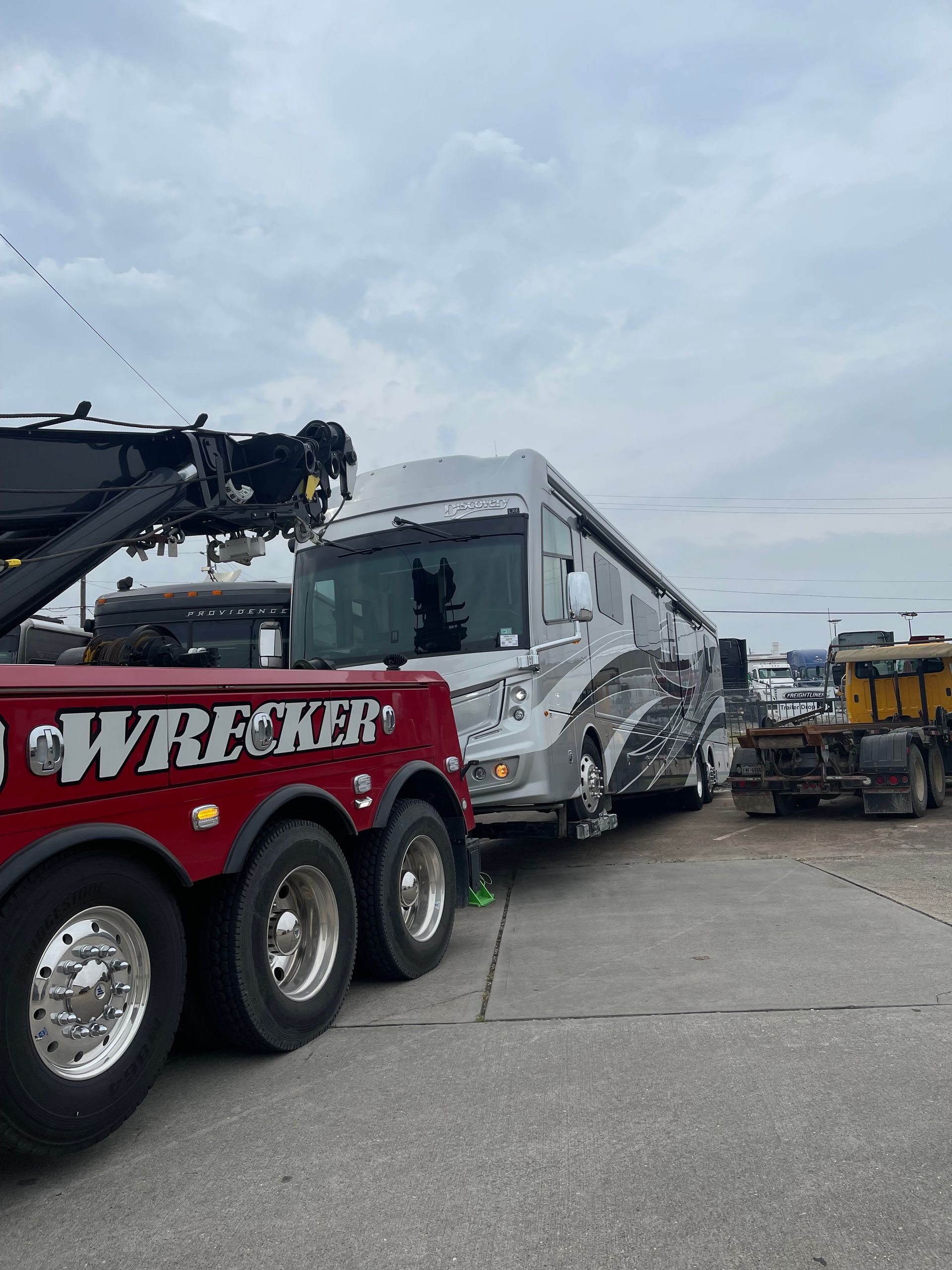 A red tow truck is towing a rv in a parking lot.