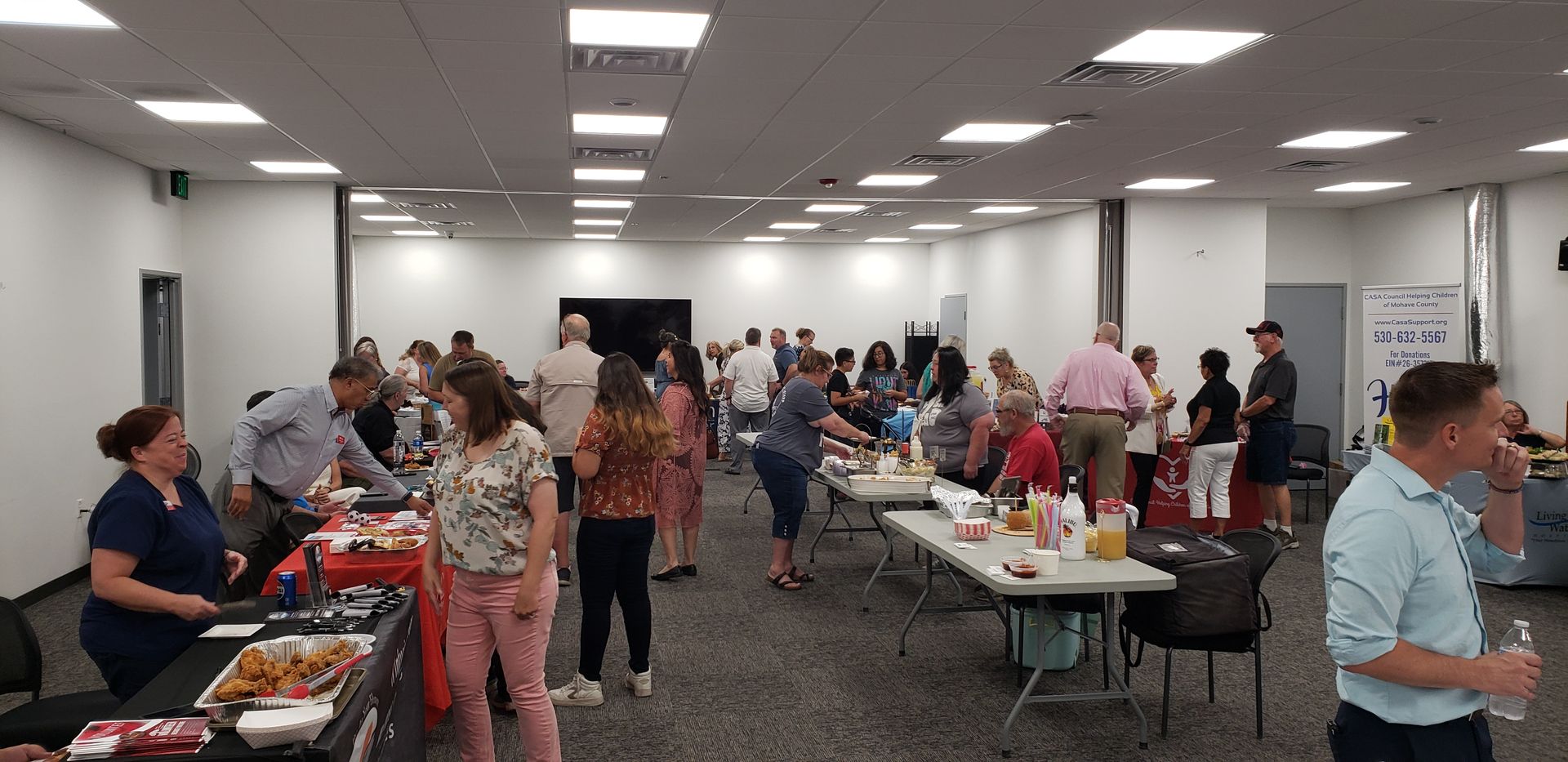 People gathered in a large room. Some are near tables with food and items. Others are talking or standing. White walls and ceiling.