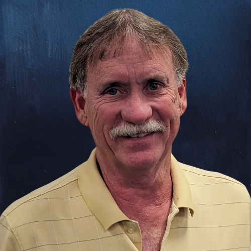 Man with a mustache wearing a yellow collared shirt smiles against a blue backdrop.