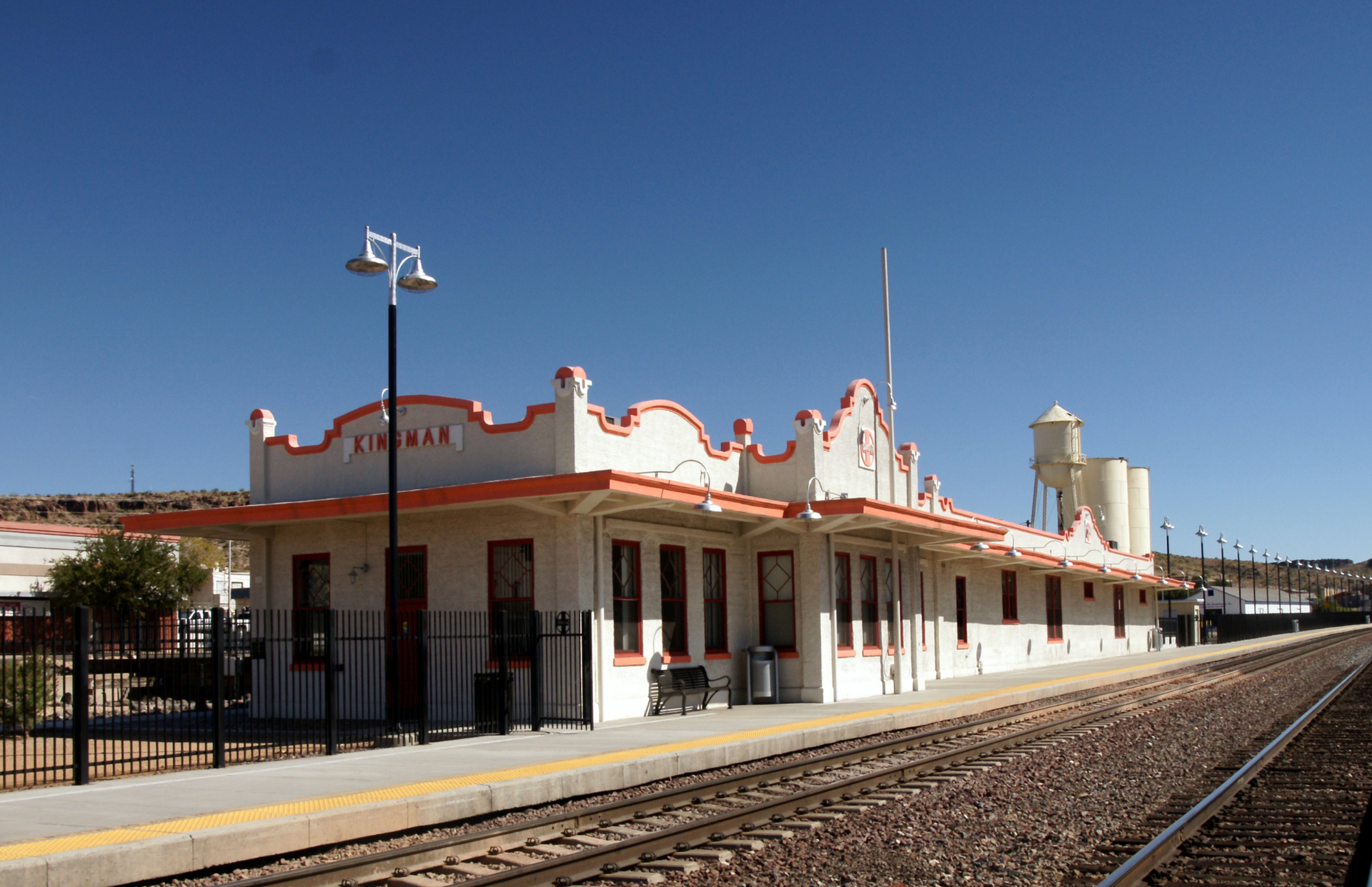 Train depot in white stucco with orange trim under a bright blue sky; railroad tracks in foreground.