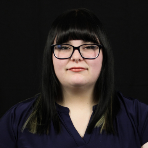 Woman with dark hair, bangs, glasses, and a nose ring, wearing a dark blue shirt, against a black backdrop.