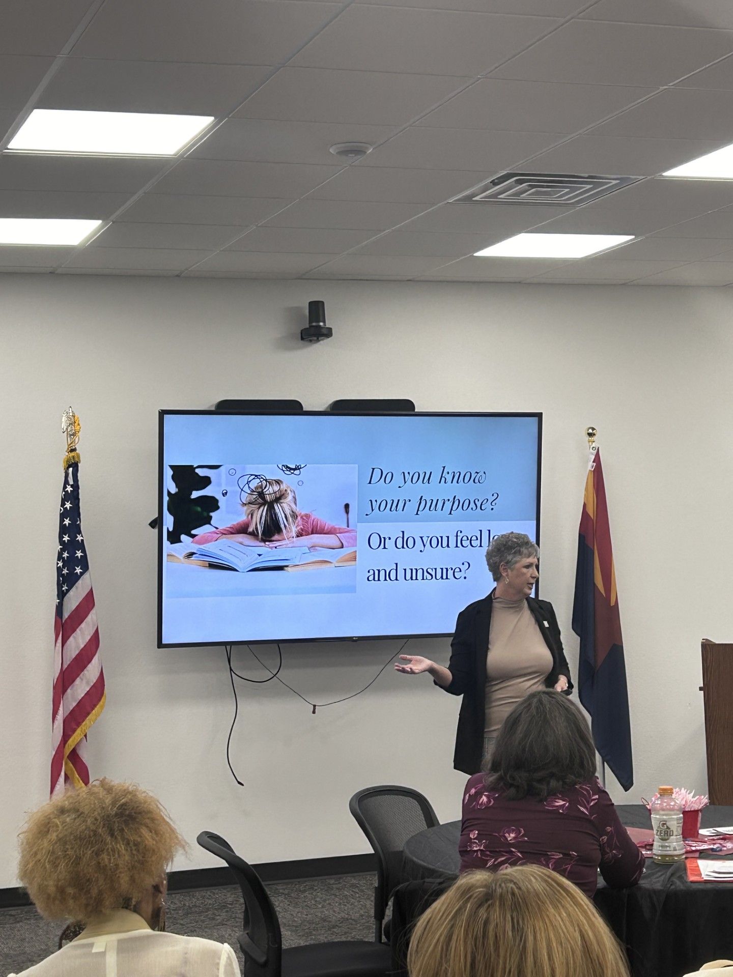 Woman giving a presentation in a room; a screen displays text and an image. An American flag and Arizona flag are present.