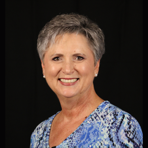 Woman with short gray hair smiles at the camera, wearing a blue patterned top, against a black backdrop.