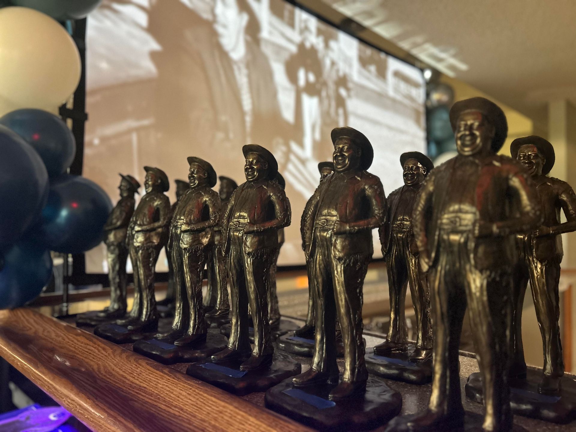 Row of bronze statuettes of a man in a hat on a wooden surface, with a projected image in the background.