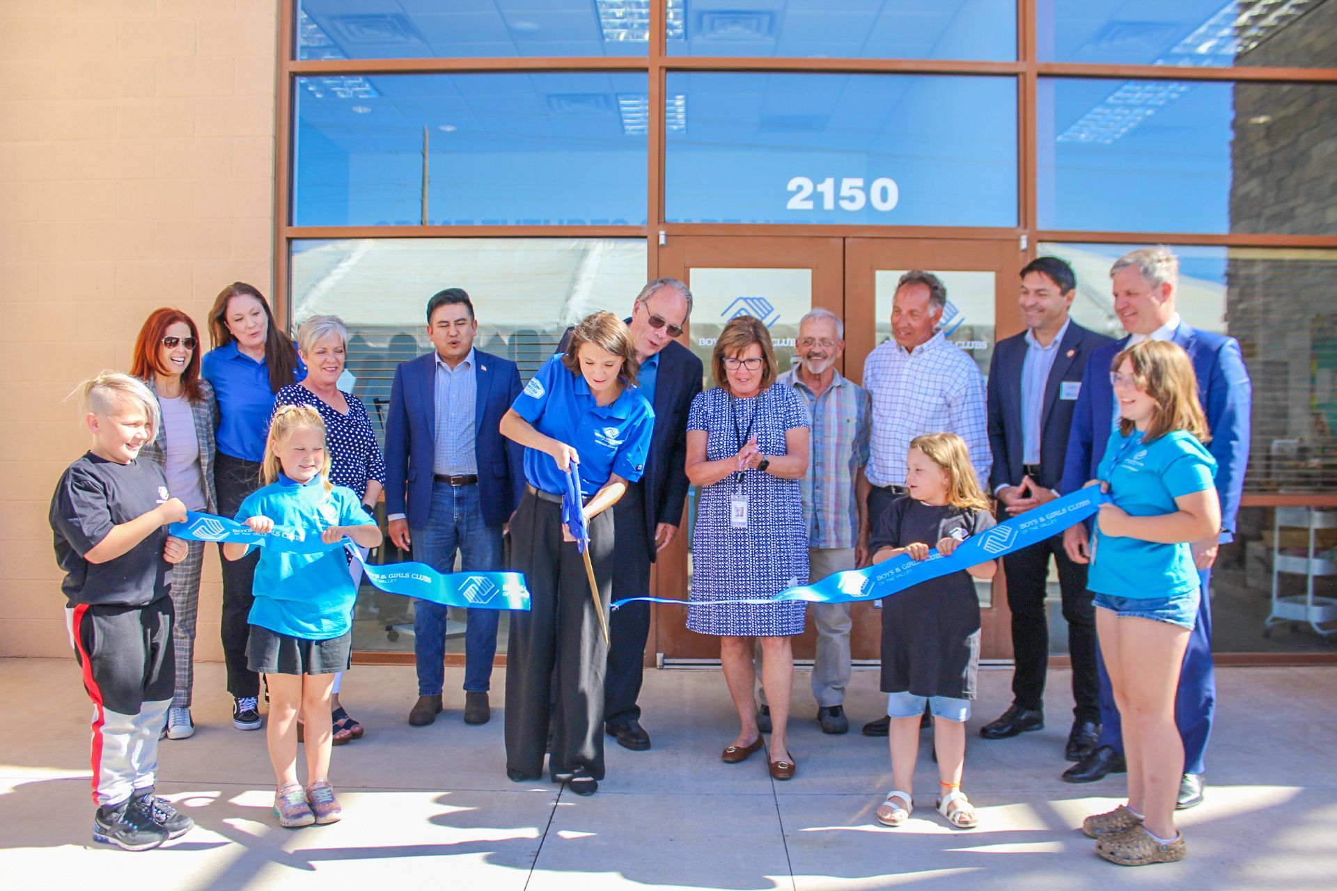 People cutting a blue ribbon in front of a building with the number 2150 on the door.