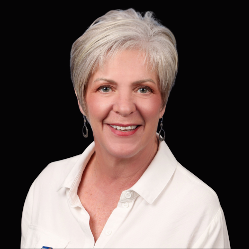 Woman with short gray hair smiling, wearing a white shirt and earrings, against a black background.
