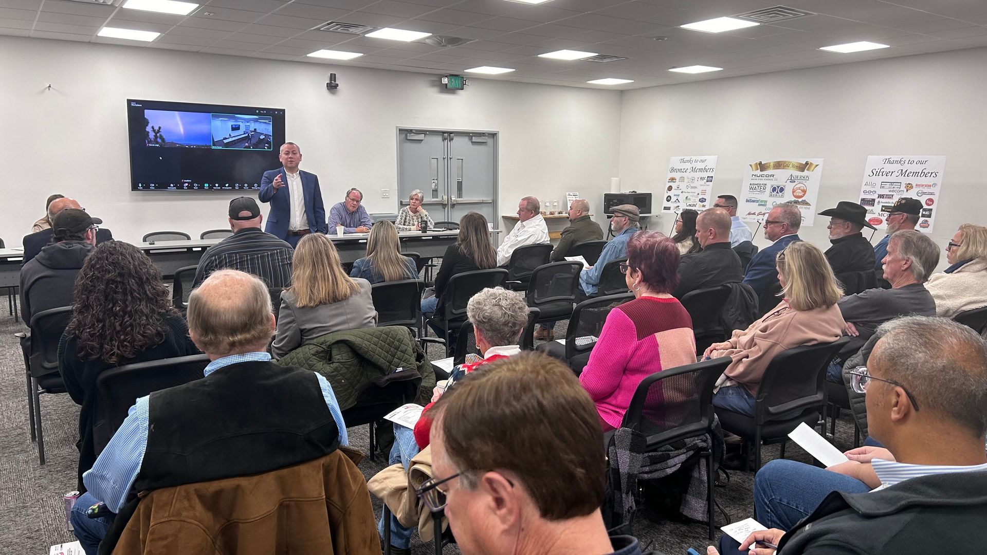 A meeting in a large room. A man speaks at a podium. People sit at tables, listening. A screen displays a video call.