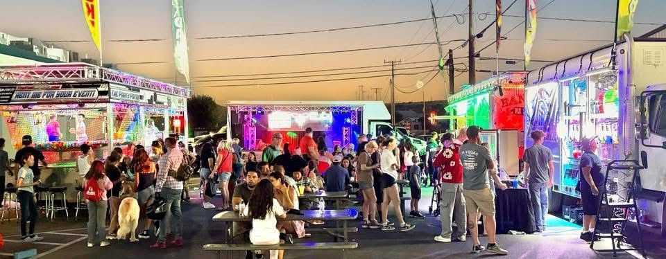 Crowd at outdoor evening food truck event with stage and colorful lights.