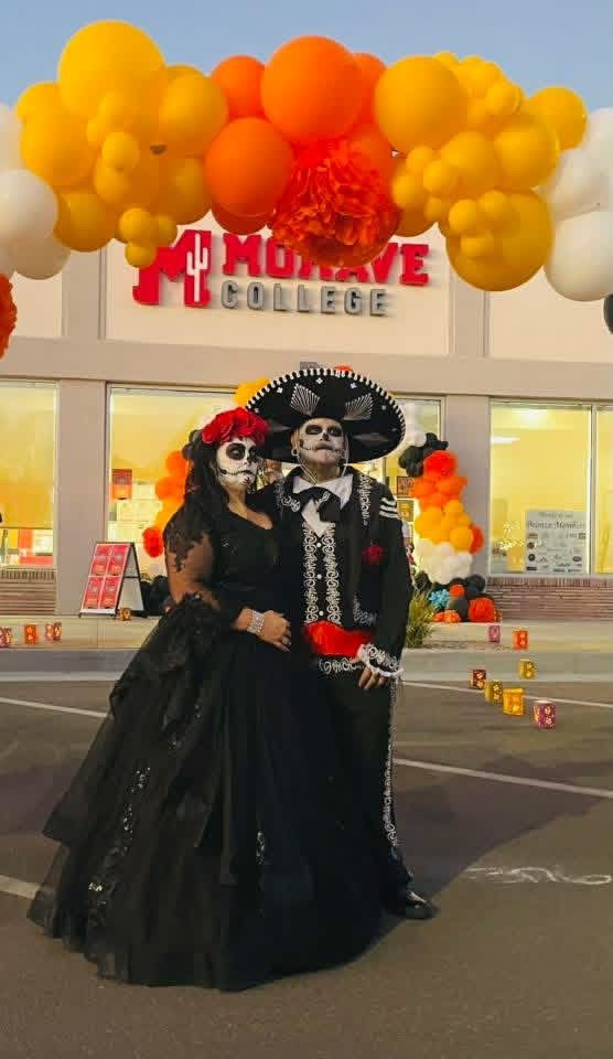Couple in Day of the Dead attire posing in front of Mohave College entrance, under balloon arch.