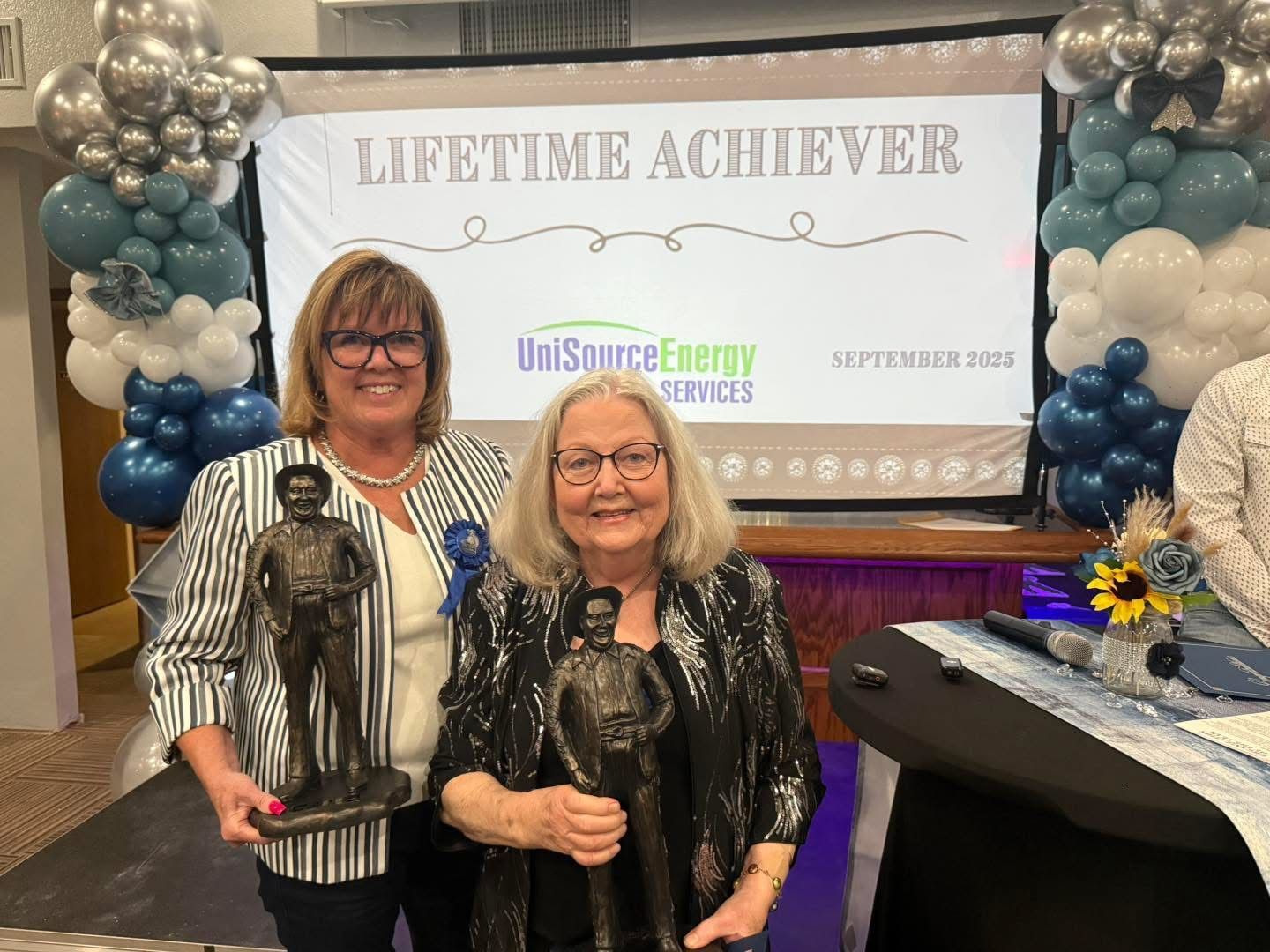 Two women holding bronze statues at an awards ceremony. A banner reads 