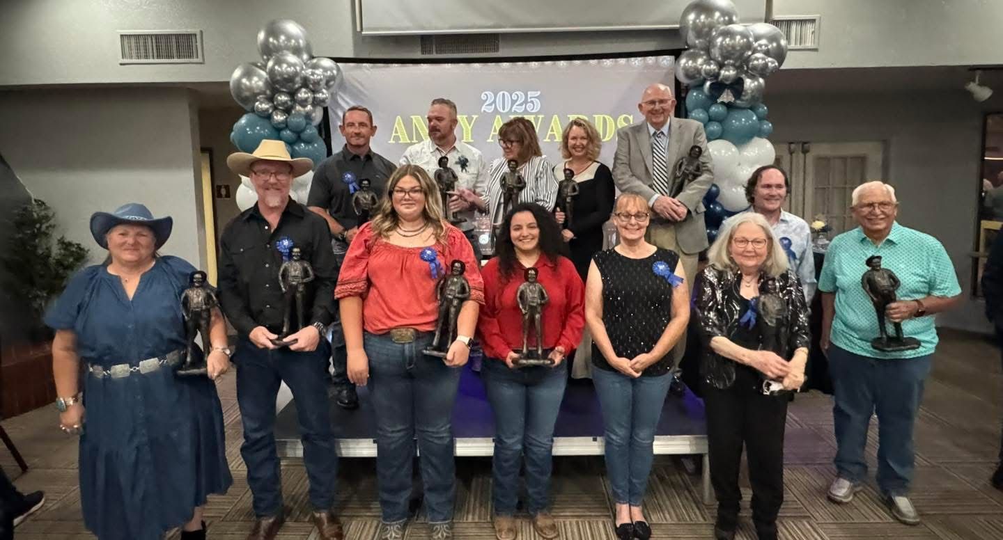 Group of people holding trophies, smiling, standing in front of a banner with balloons.