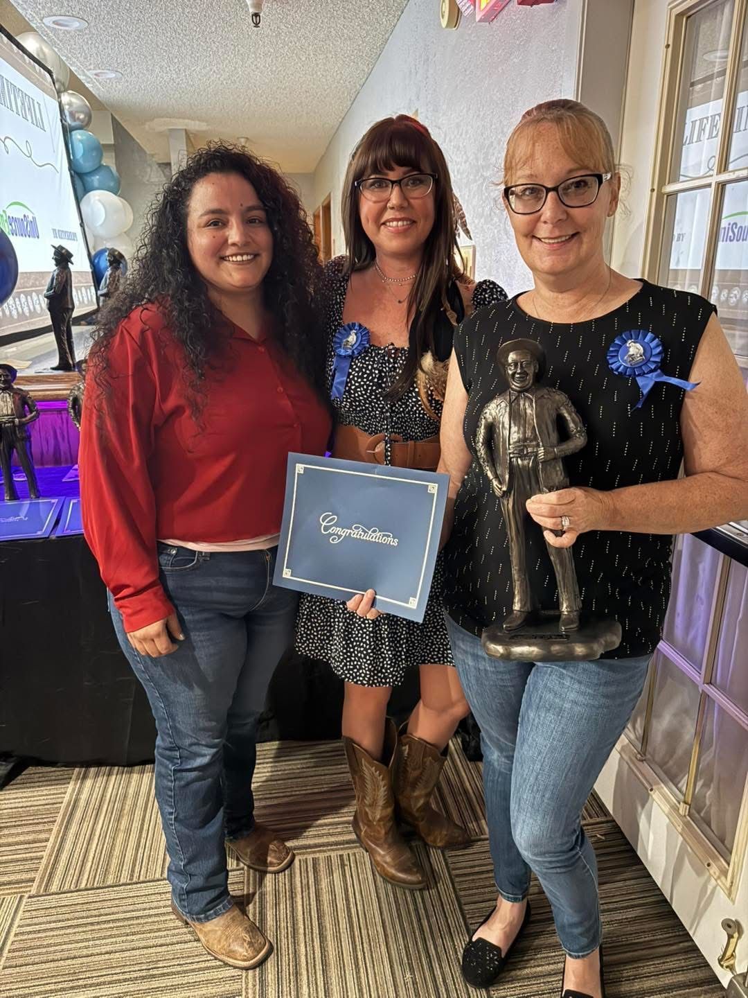 Three women at an event, one holding an award and a certificate. Smiling and posing near a door and blue decorations.