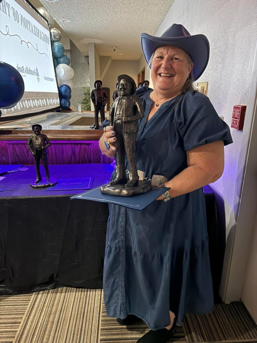 Woman in a blue dress and hat, holding a statue. Other similar statues are on a table.