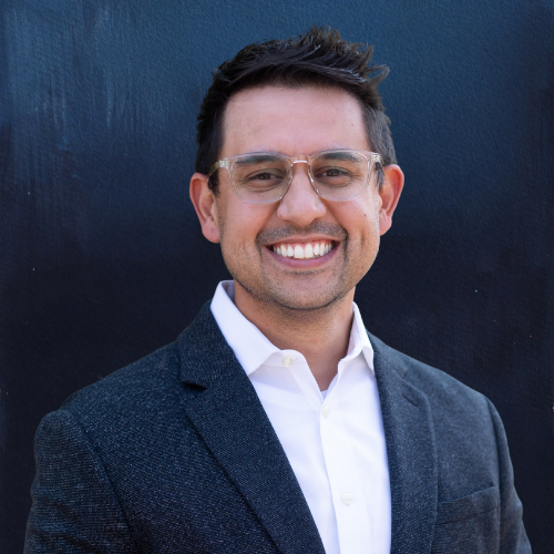 Man with glasses smiles, wearing a suit jacket and white shirt against a dark background.