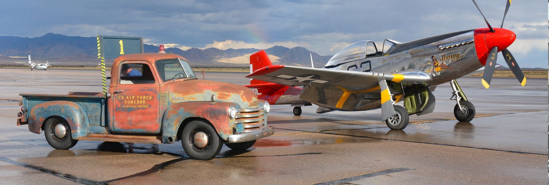 Rusty truck parked next to a P-51 Mustang airplane on an airfield. Mountains in the background.