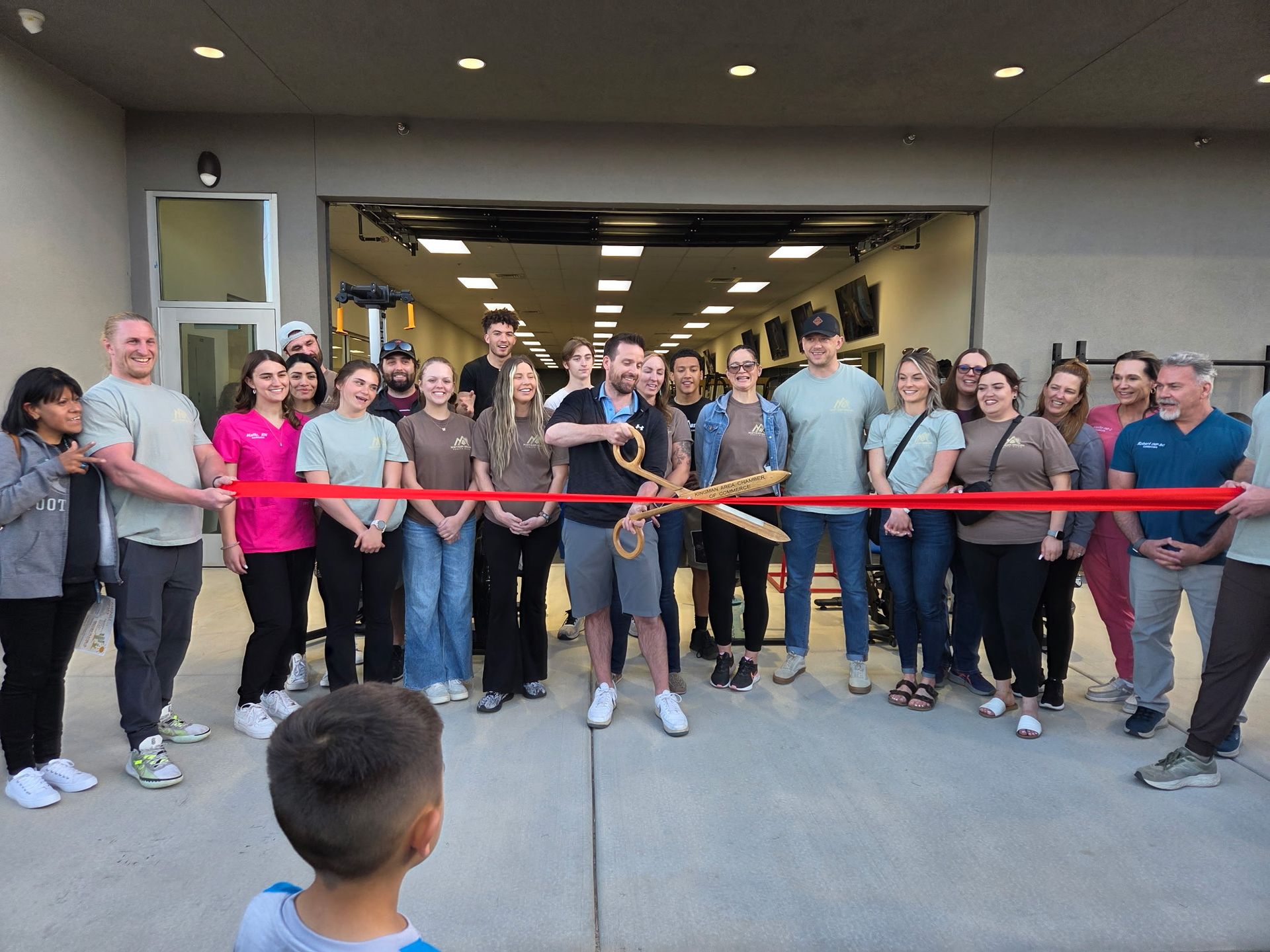 Group of people cutting a red ribbon with large gold scissors at an outdoor event.