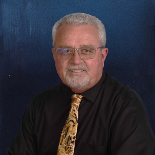 Man with glasses and goatee wearing a black shirt and patterned tie, smiling against a blue background.