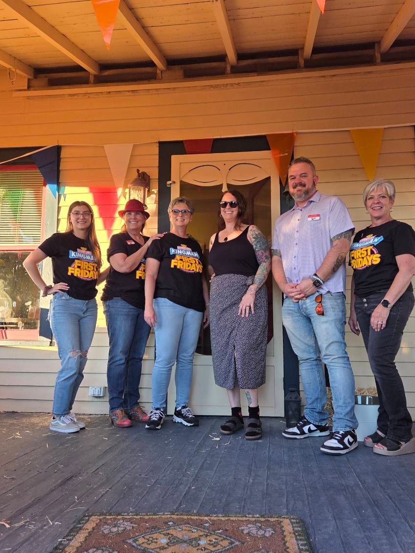 Six people stand on a porch. They wear matching t-shirts. A banner hangs above the doorway.