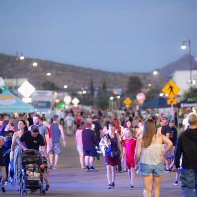Crowd of people walking down a street at dusk. Street vendors and mountain in background.