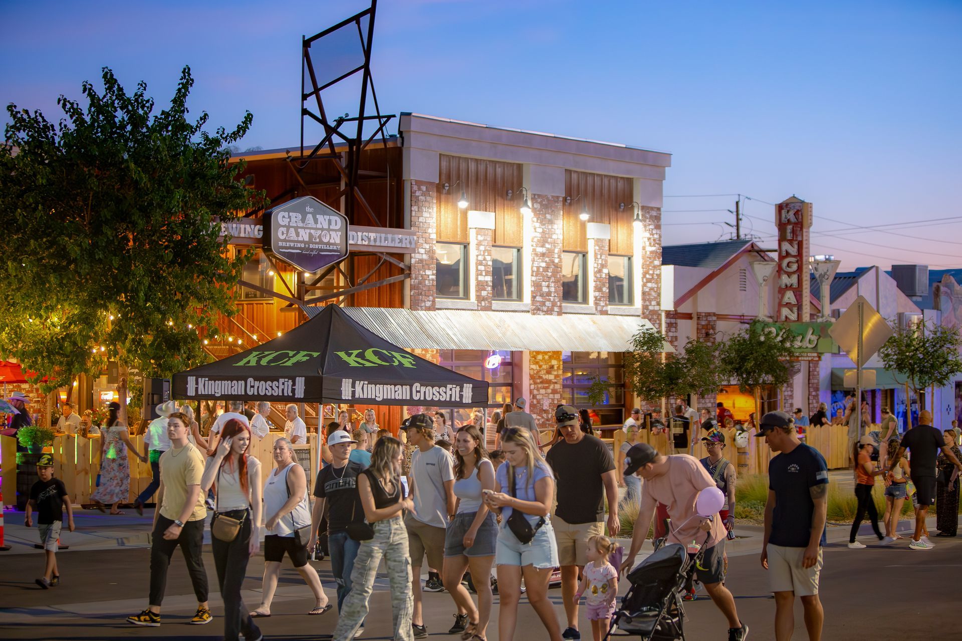 Crowd of people walking outdoors at dusk, near lit buildings with awnings and signage.