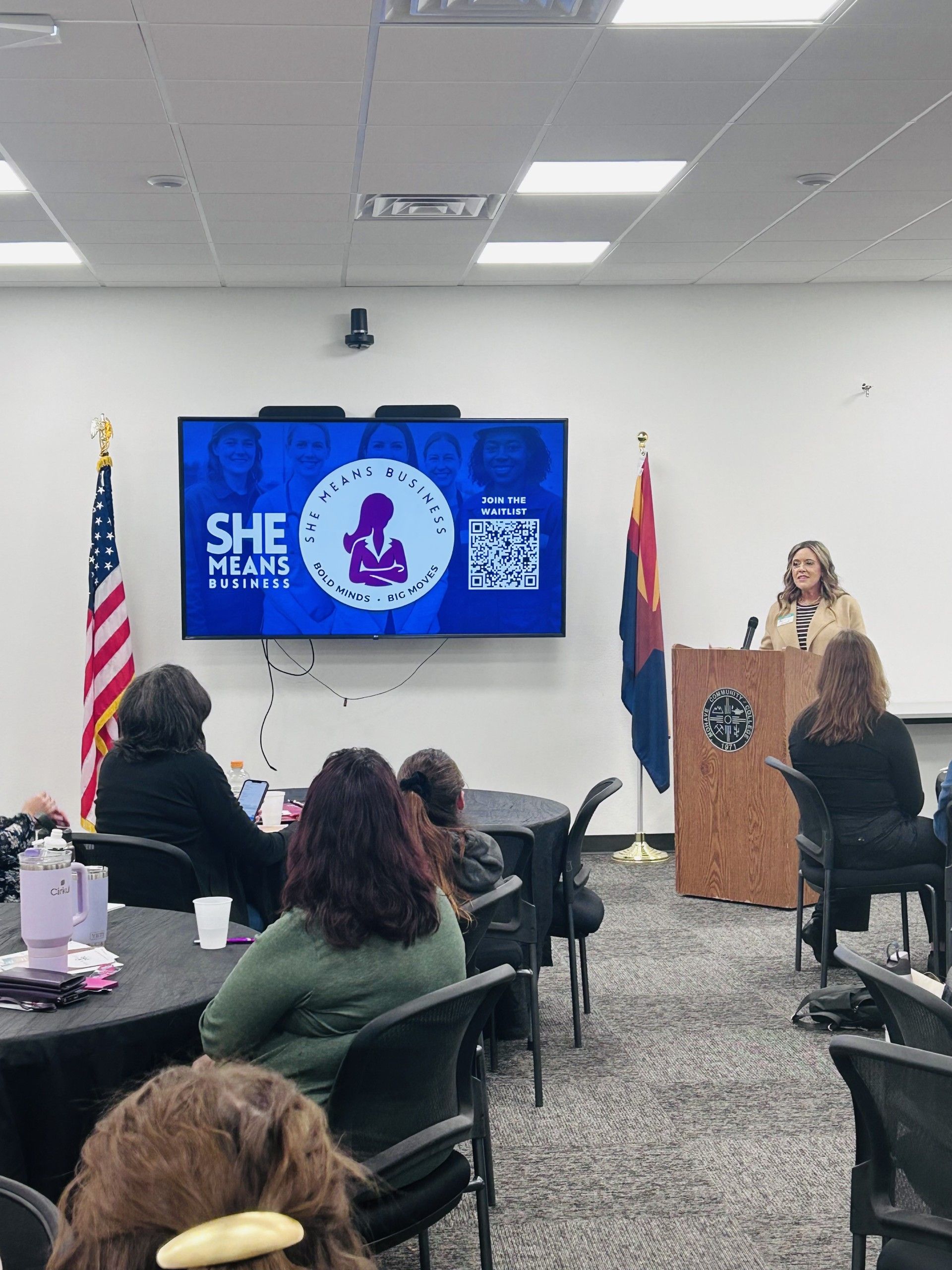 Woman speaking at a podium in a room with an audience; presentation screen shows