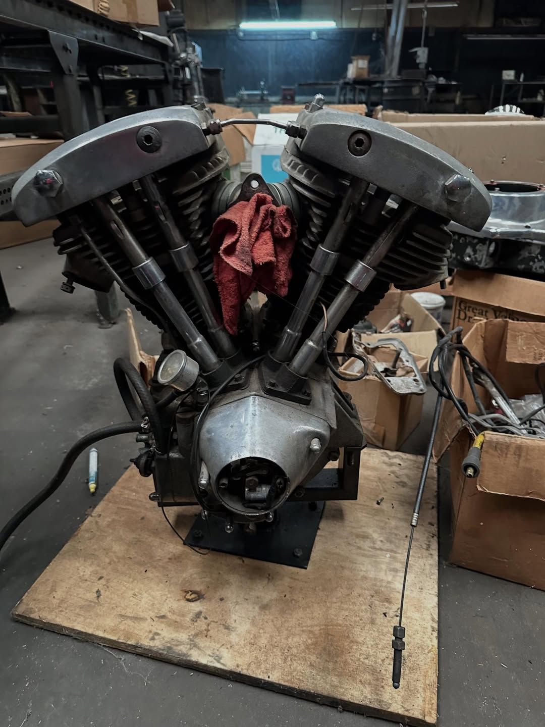 V-twin motorcycle engine on a wooden platform inside a workshop, with metal components and a red rag.