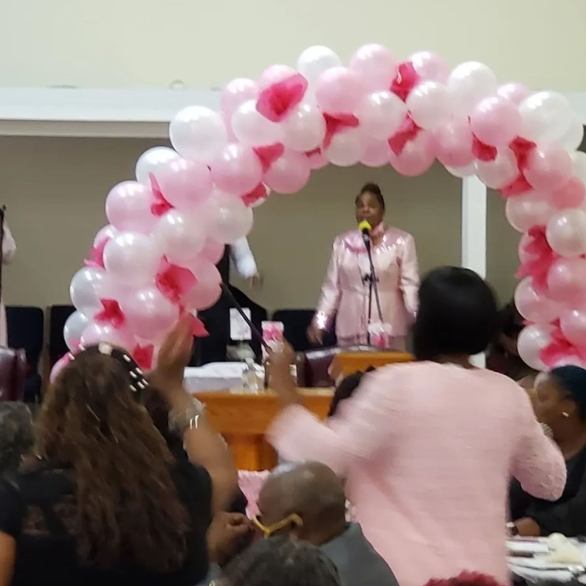 A woman stands at a podium surrounded by pink and white balloons