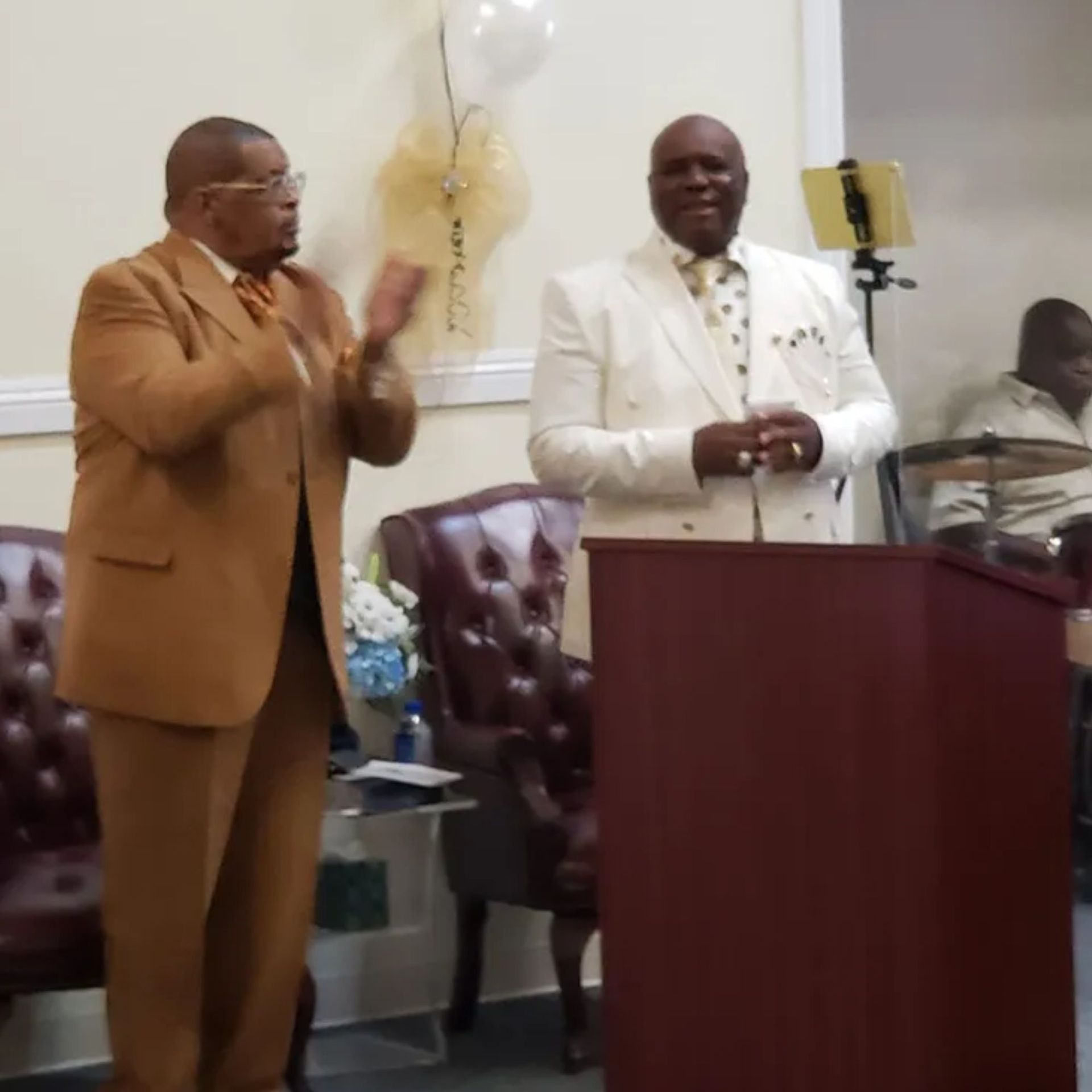 A man in a brown suit stands next to a man in a white suit at a podium