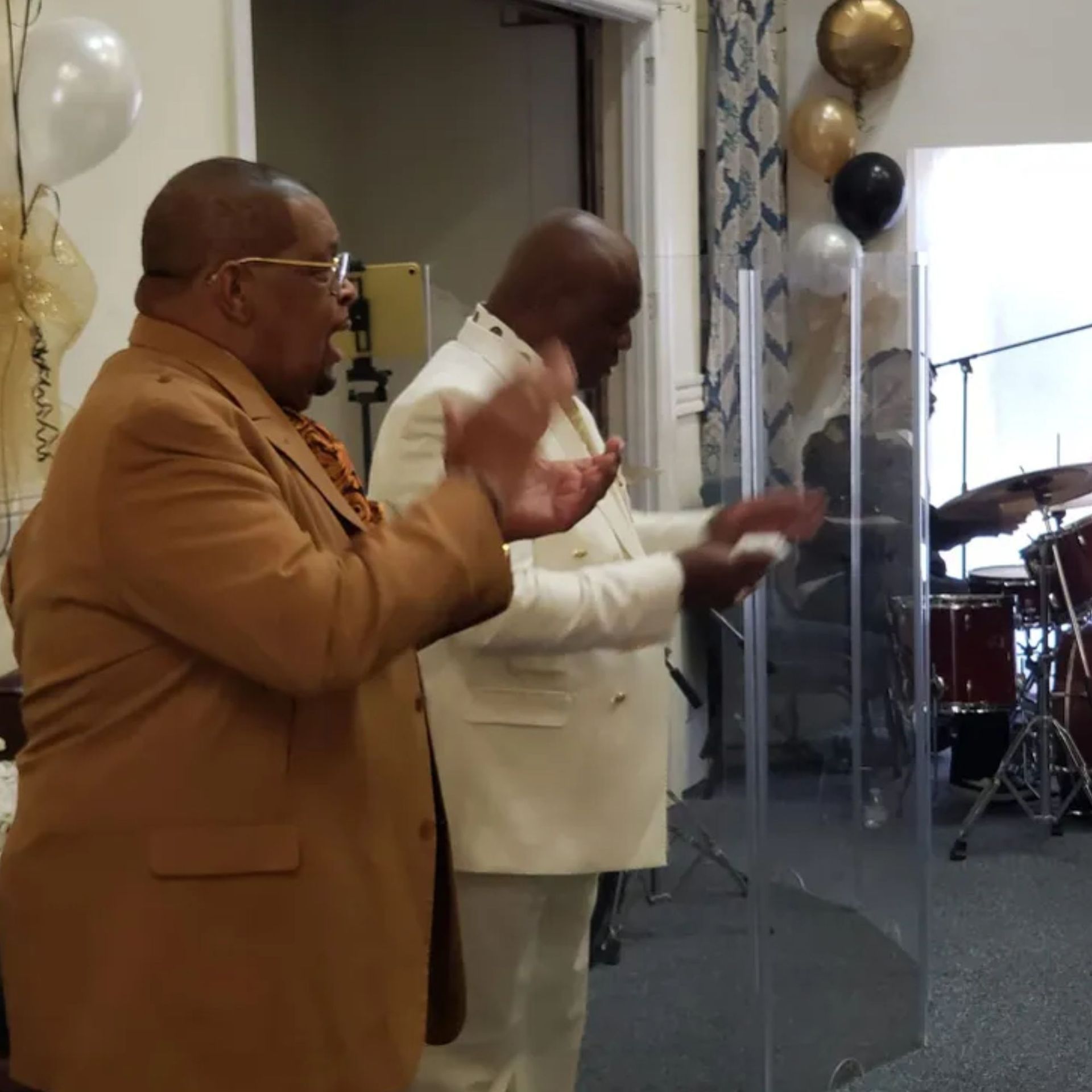 A man in a brown suit stands next to another man in a white suit