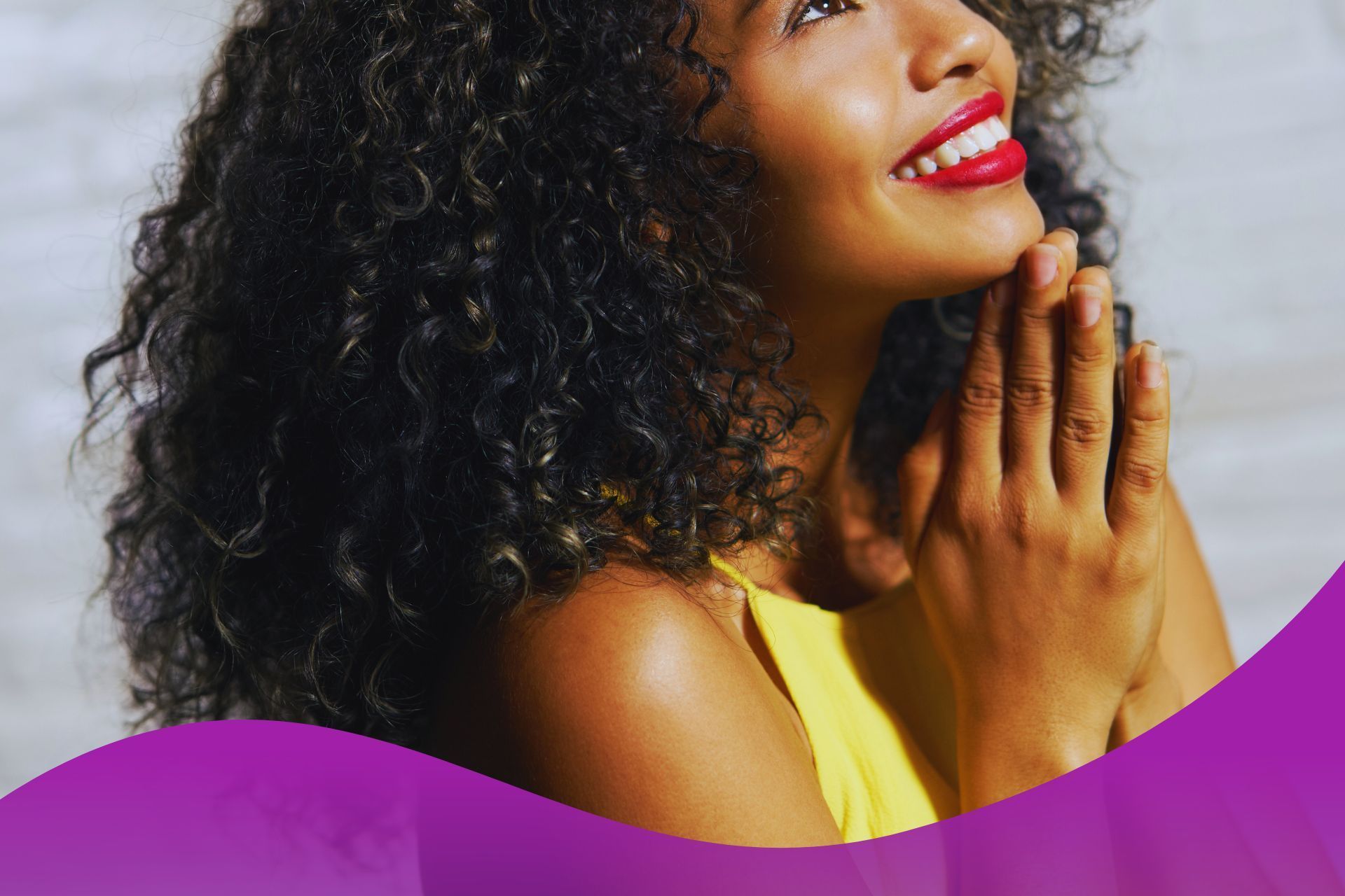 A woman with curly hair is smiling with her hands folded in prayer.