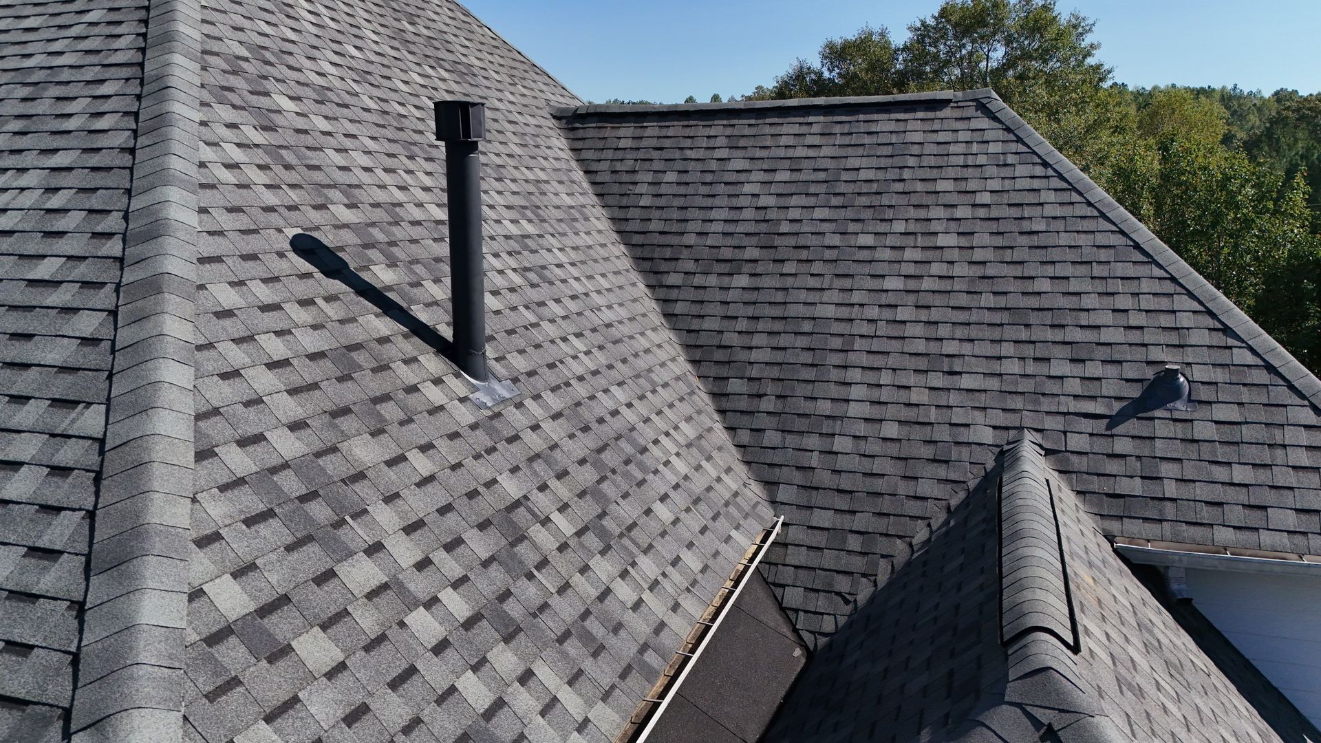 An aerial view of a dark grey shingled roof with a black chimney pipe and a valley.