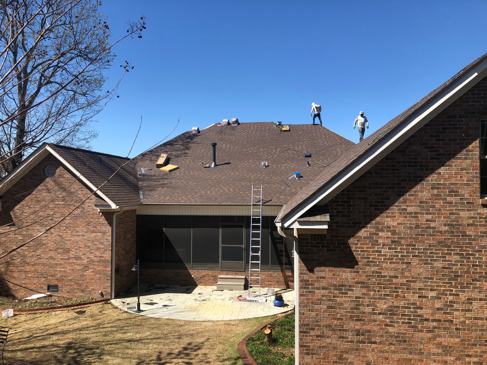 Two workers stand on the shingled roof of a brick home during a construction project, with a ladder leaning against it.